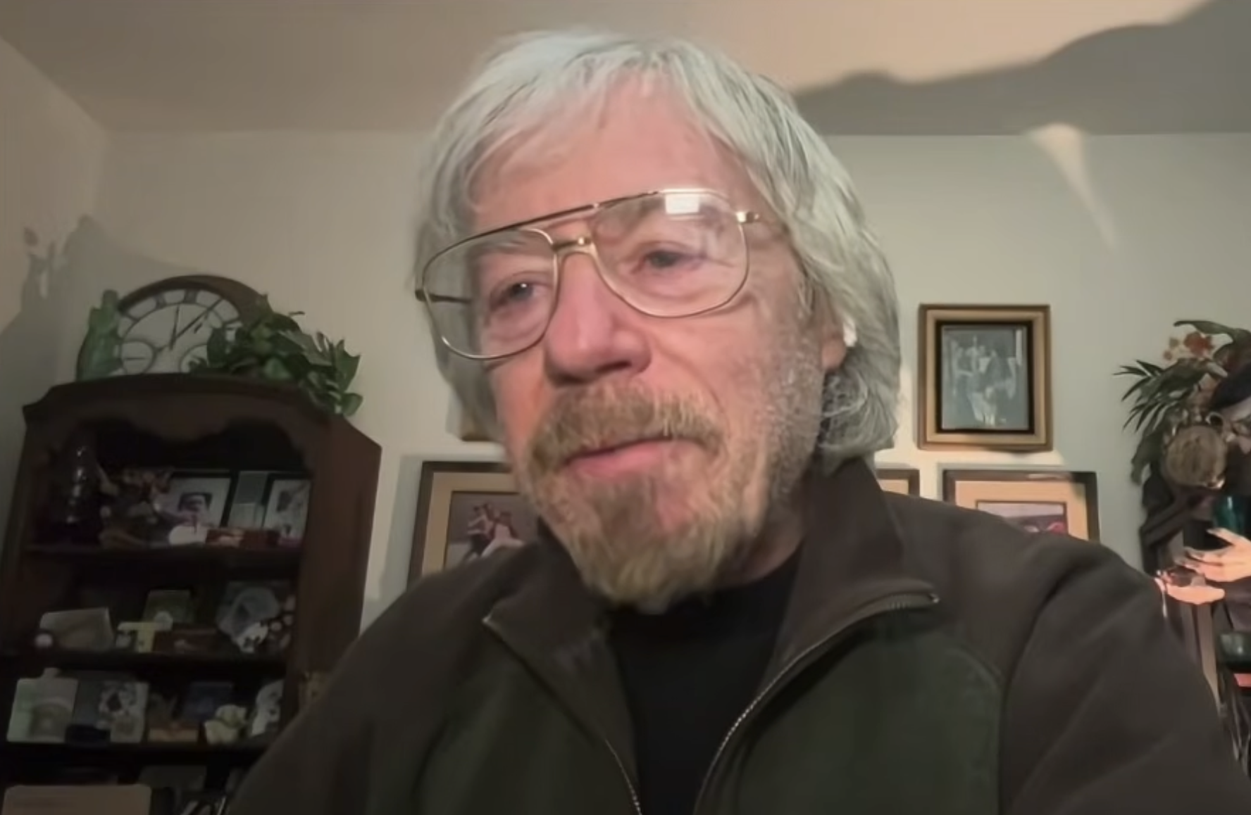 An older man with gray hair, beard, and large glasses is sitting indoors. He is wearing a dark jacket. Behind him are framed photos on the wall, a shelf with plants, and other decorative items.