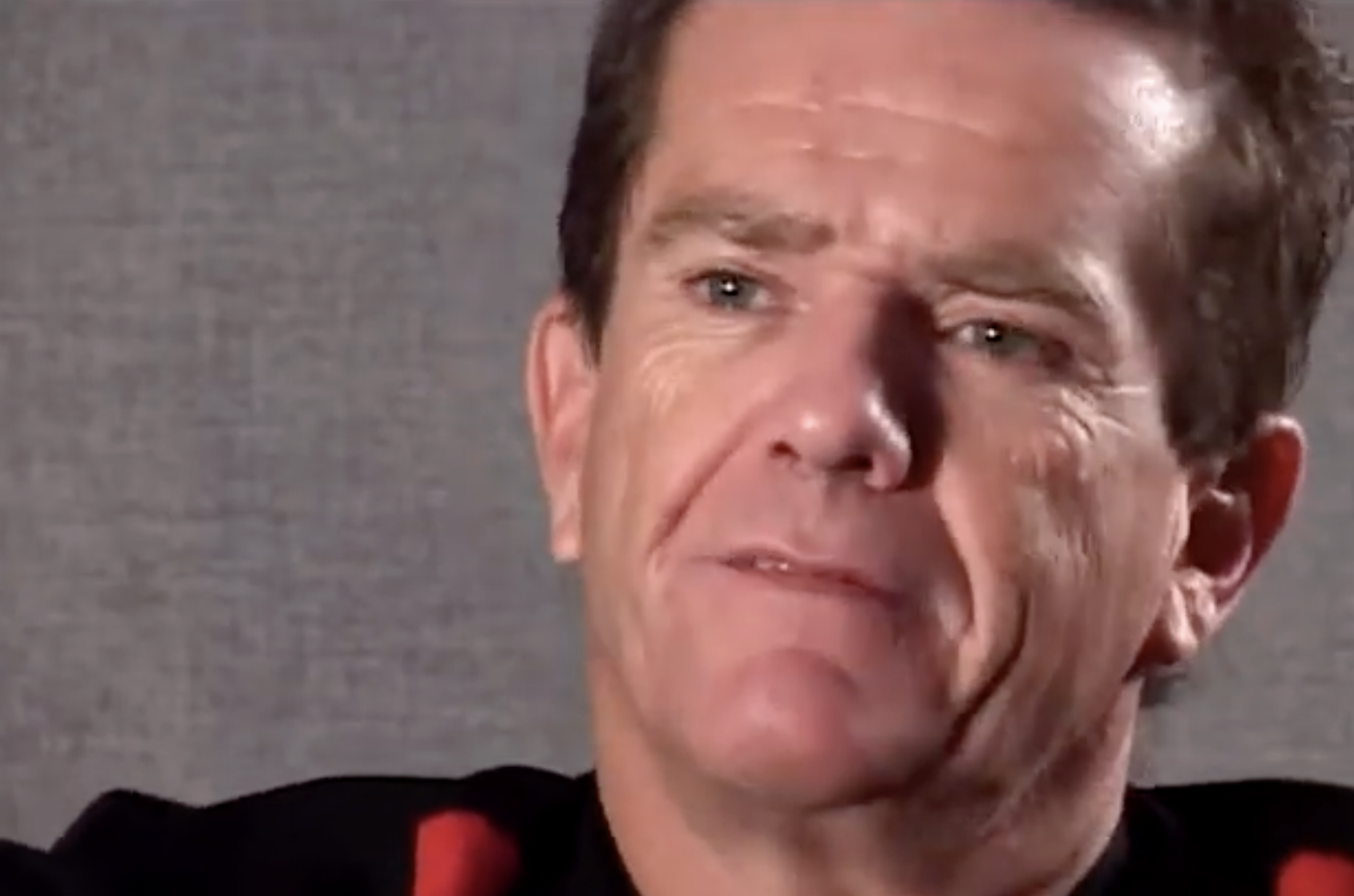 A middle-aged man with short brown hair and a slight frown looks off-camera. He is wearing a black shirt with red accents and is posed against a plain grey background.