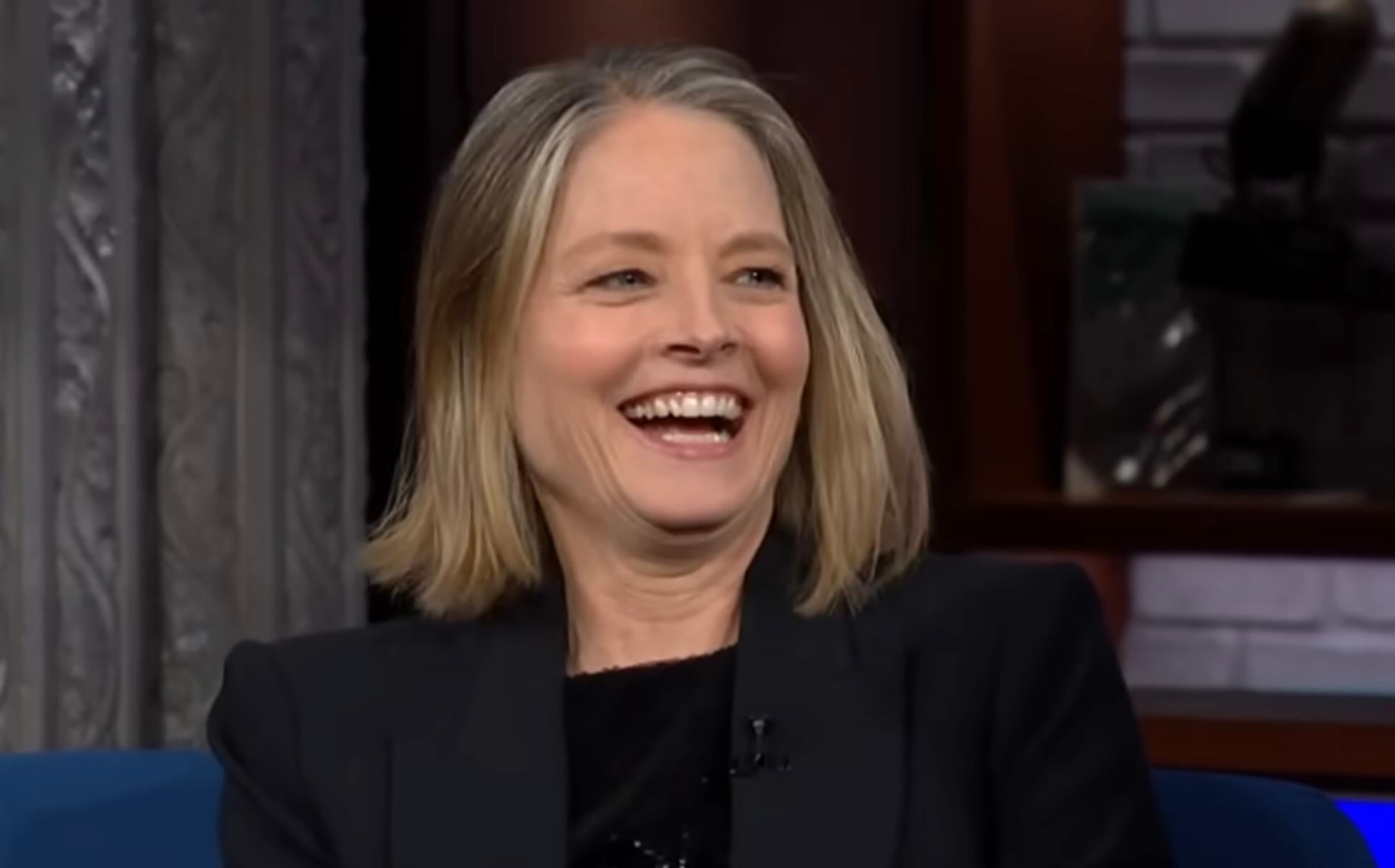 A woman with shoulder-length blonde hair is smiling and laughing while sitting indoors, wearing a black blazer and a black top. The background is blurred, showing shelves and part of a curtain.