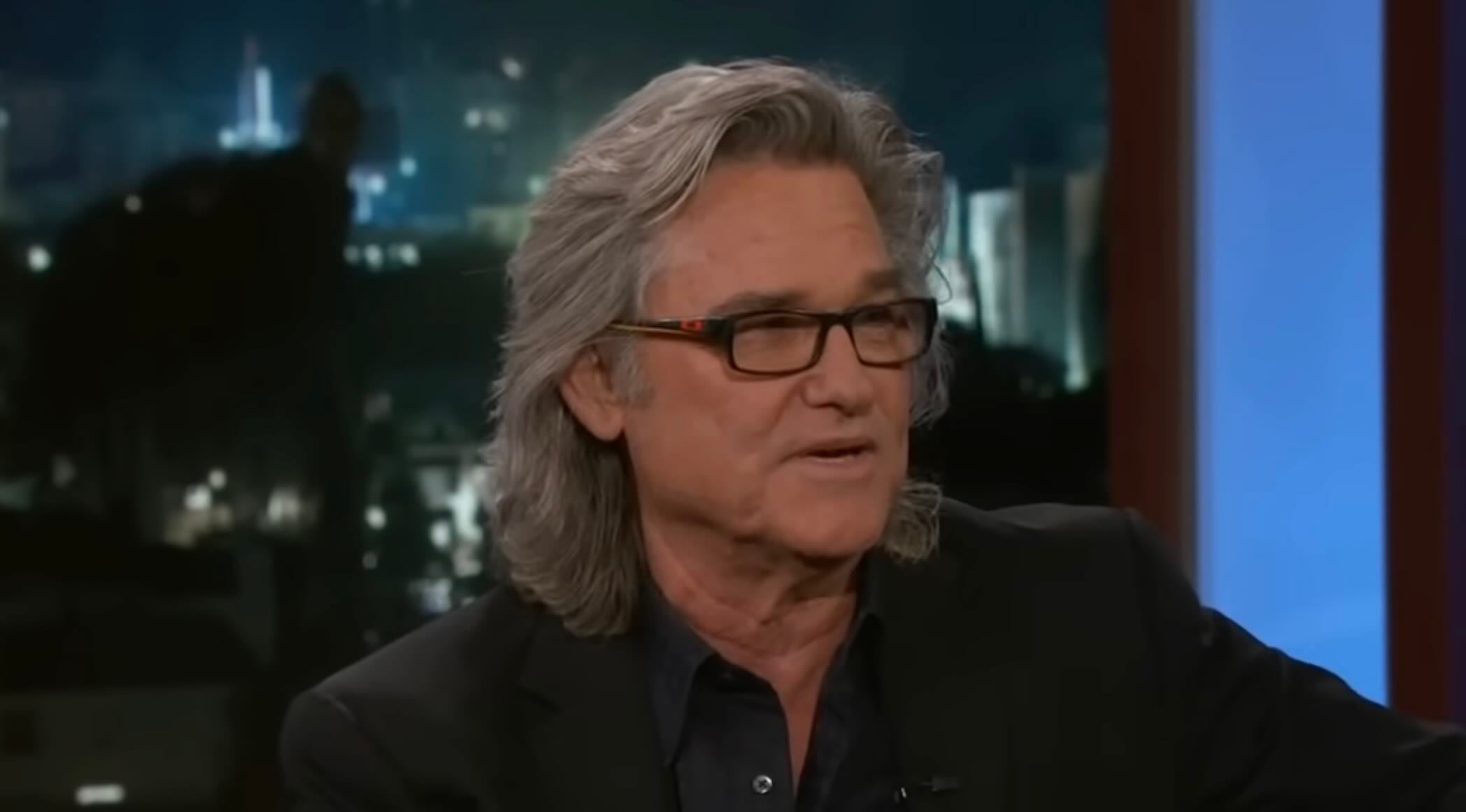 A man with gray, shoulder-length hair and glasses is wearing a dark blazer and speaking during a TV interview, with a blurred nighttime cityscape in the background.