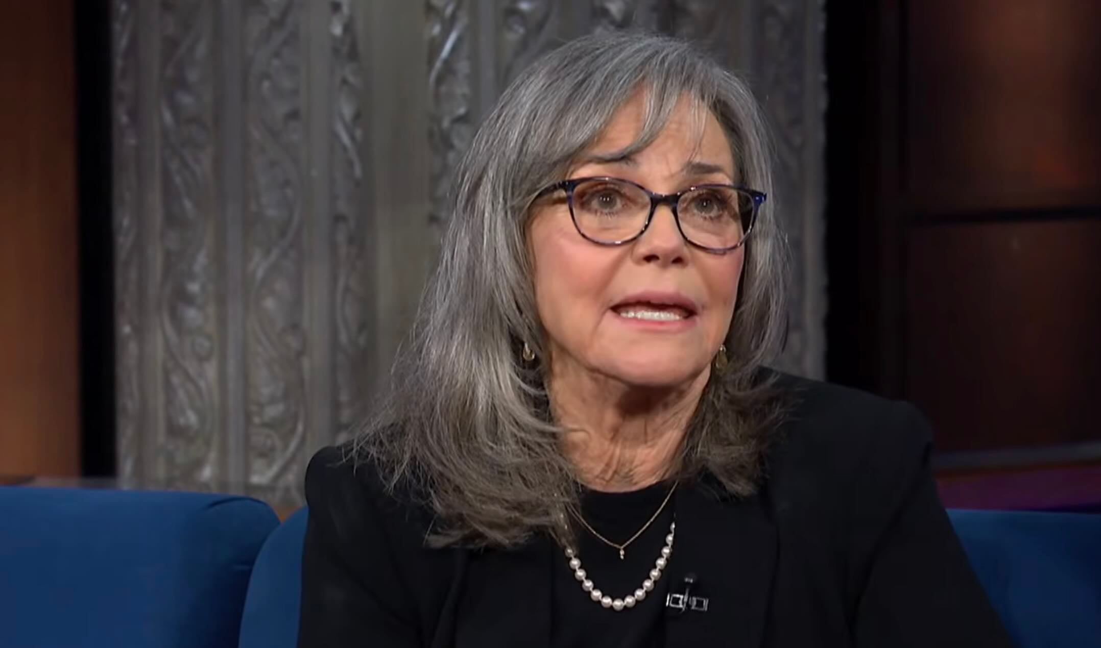 An older woman with gray hair and glasses, wearing a black outfit and a pearl necklace, sits on a blue couch during a TV interview. Ornate metallic decor is visible in the background.