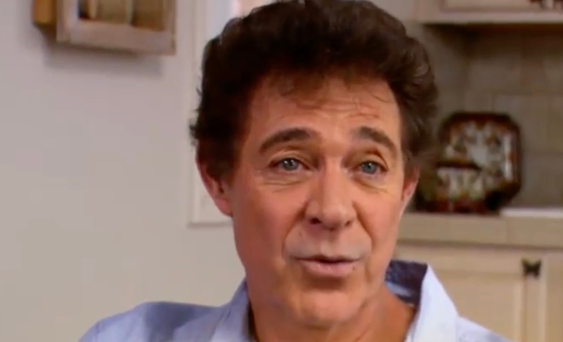 A middle-aged man with curly brown hair wearing a light blue shirt is sitting indoors, speaking or mid-sentence. The background shows a kitchen with white cabinets and a decorative plate.