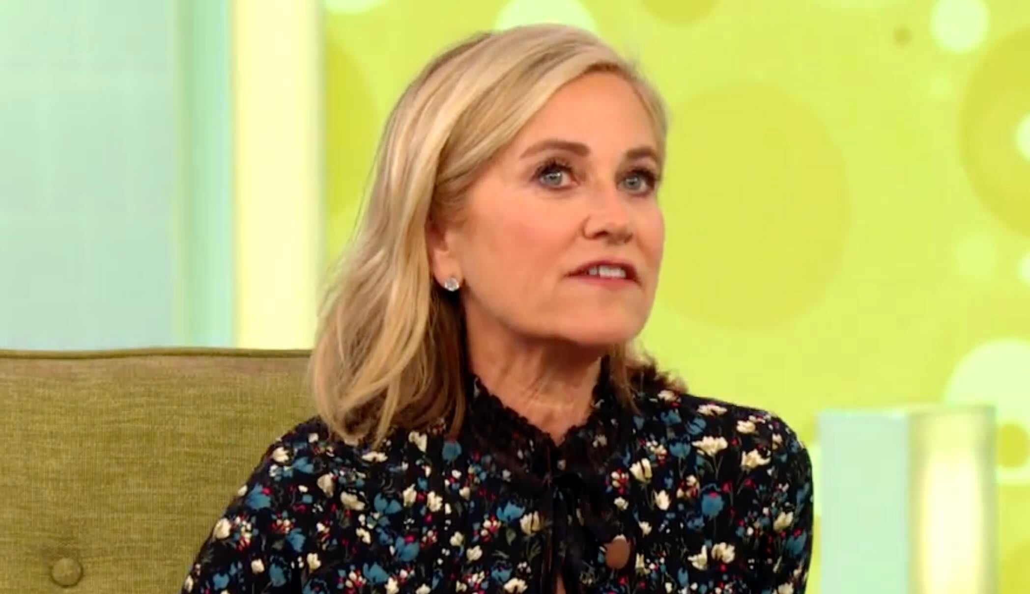 A woman with shoulder-length blonde hair sits on a green chair, wearing a black floral blouse, against a yellow background with circular patterns. She appears to be speaking or engaged in conversation.