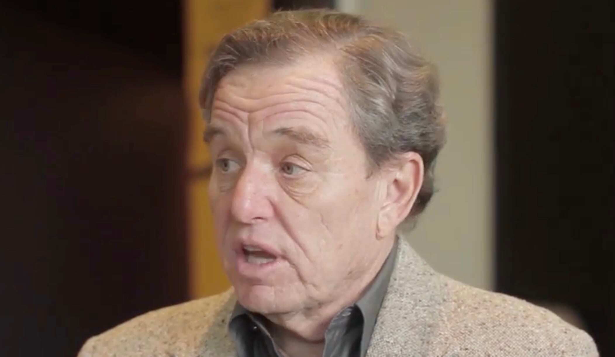 An older man with gray hair, wearing a gray shirt and a beige blazer, is speaking with his mouth slightly open. He appears to be in mid-conversation, with a neutral background behind him.