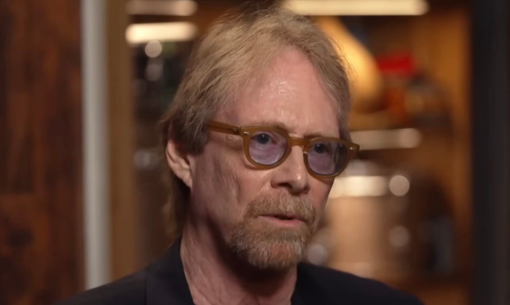 A middle-aged man with light brown hair, a short beard, and light brown glasses is wearing a dark jacket and looking slightly to the side in a softly lit indoor setting.