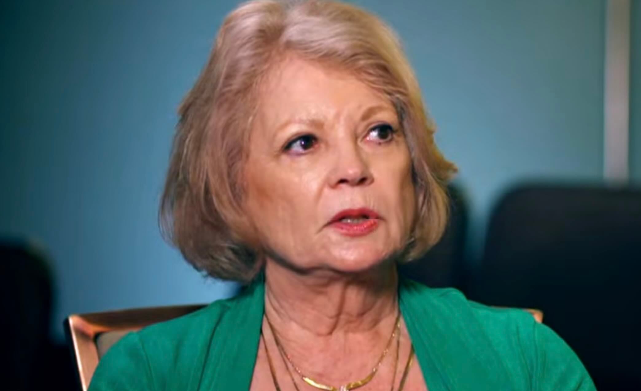 An older woman with short blonde hair and light skin, wearing a green top and a gold necklace, looks slightly to the side with a serious expression against a blurred blue background.