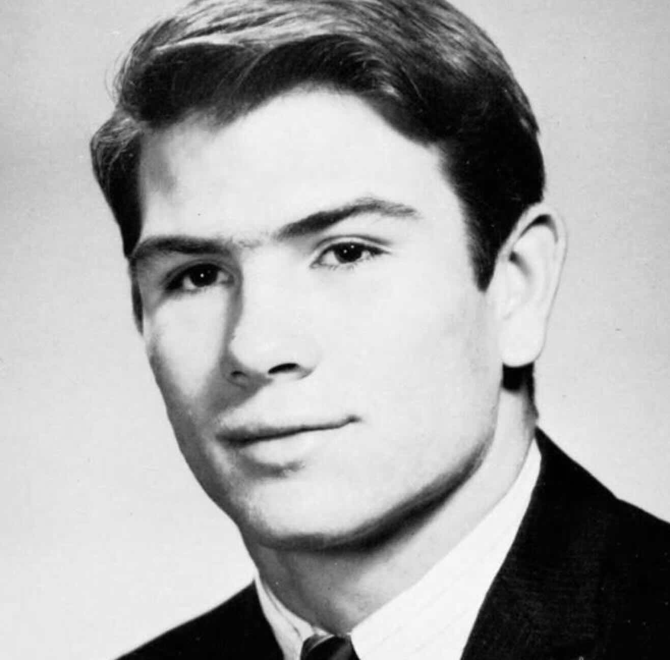 Black and white portrait of a young man with short, neatly combed hair, wearing a suit jacket, dress shirt, and tie, looking slightly past the camera with a neutral expression.