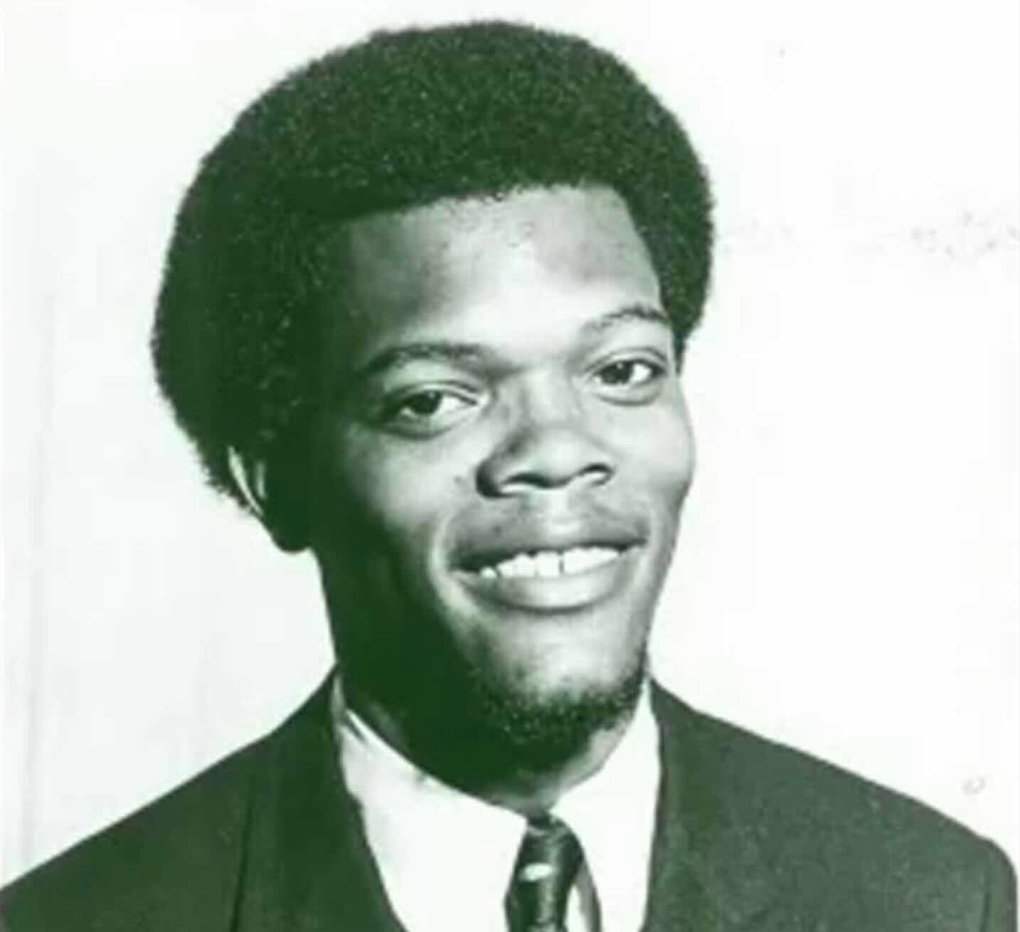 A young man with a short afro hairstyle and a goatee smiles at the camera. He is wearing a suit, white shirt, and patterned tie. The photo is black and white with a plain background.