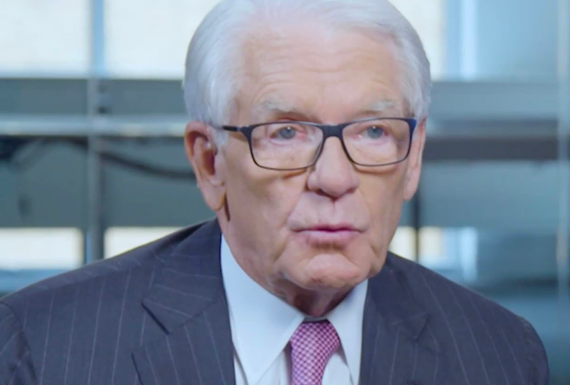 An older man with white hair and glasses, wearing a dark pinstripe suit, white shirt, and light pink tie, is speaking indoors with a blurred office background.