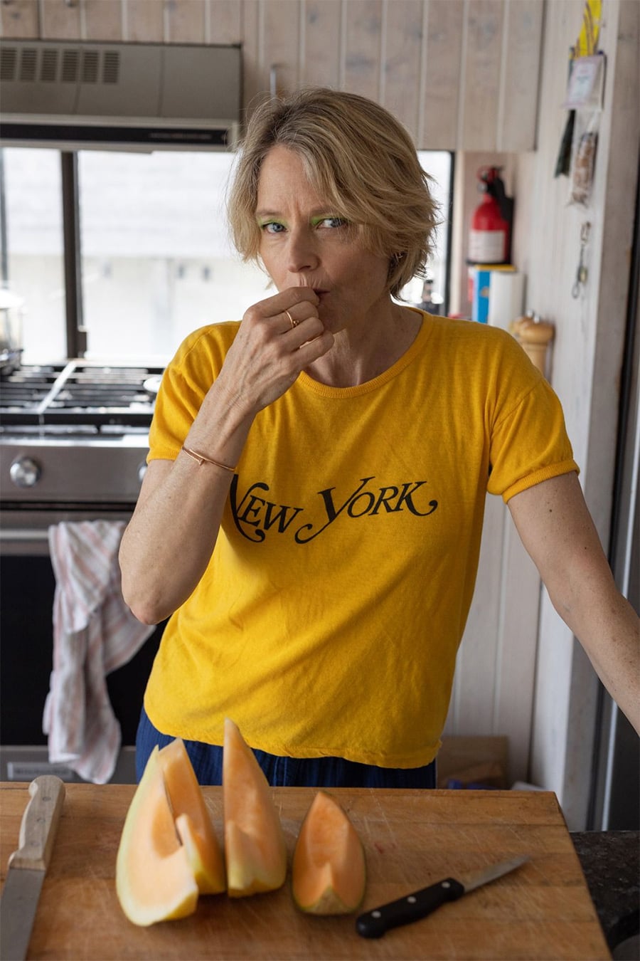 A woman in a yellow "New York" t-shirt stands in a kitchen, holding a piece of cantaloupe to her mouth. On the counter in front of her are sliced pieces of cantaloupe and a knife.