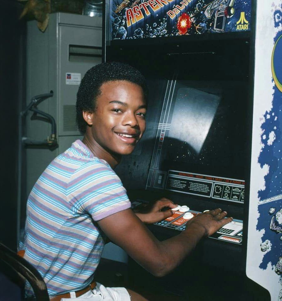 A smiling teen wearing a striped shirt plays an Asteroids Deluxe arcade game, using the controls with both hands.