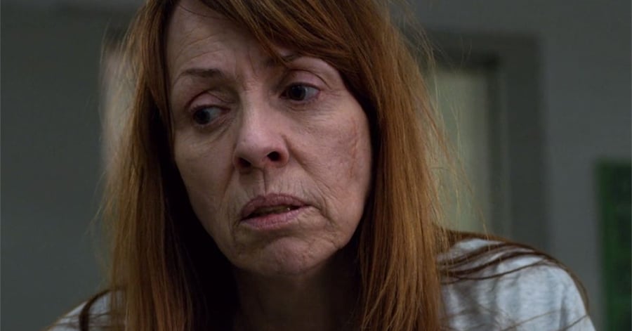 A woman with long reddish-brown hair and a serious, tired expression looks downward. She is wearing a light-colored shirt and is indoors with a blurred background.