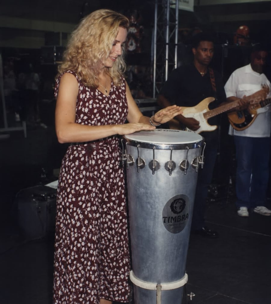 A woman in a patterned dress plays a tall drum on stage, with two musicians playing guitar and bass in the background.