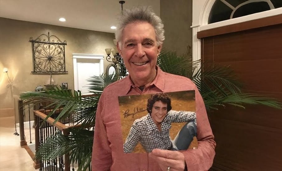 An older man in a red shirt stands indoors, smiling and holding a photo of his younger self sitting outdoors on grass, wearing a plaid shirt and smiling. The room has plants, a staircase, and warm lighting.