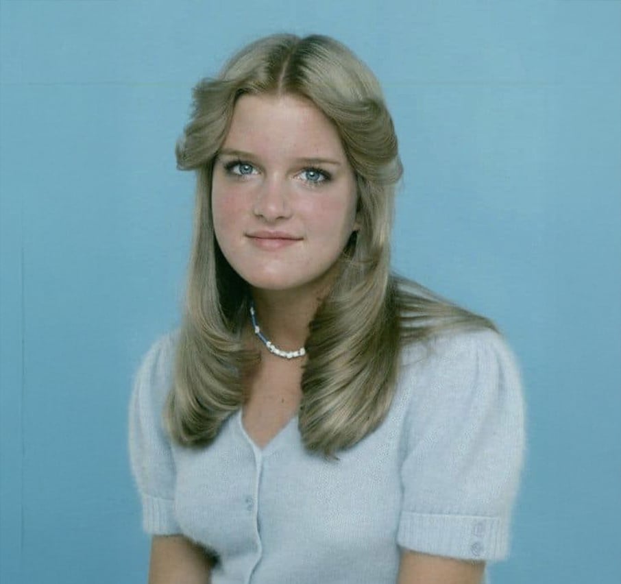 A young woman with long, blond hair styled in soft waves, wearing a light blue cardigan and a pearl necklace, sits in front of a plain blue background, looking at the camera with a slight smile.