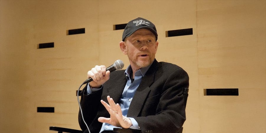 A man wearing a black baseball cap, blazer, and blue shirt holds a microphone and gestures with his hand while seated against a light wooden wall with small black rectangular accents.