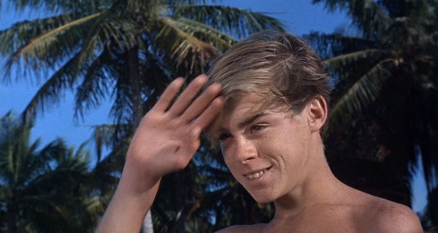 A young, shirtless boy with blond hair smiles and raises his hand, as if waving or shielding his eyes. Palm trees and a clear blue sky are in the background.