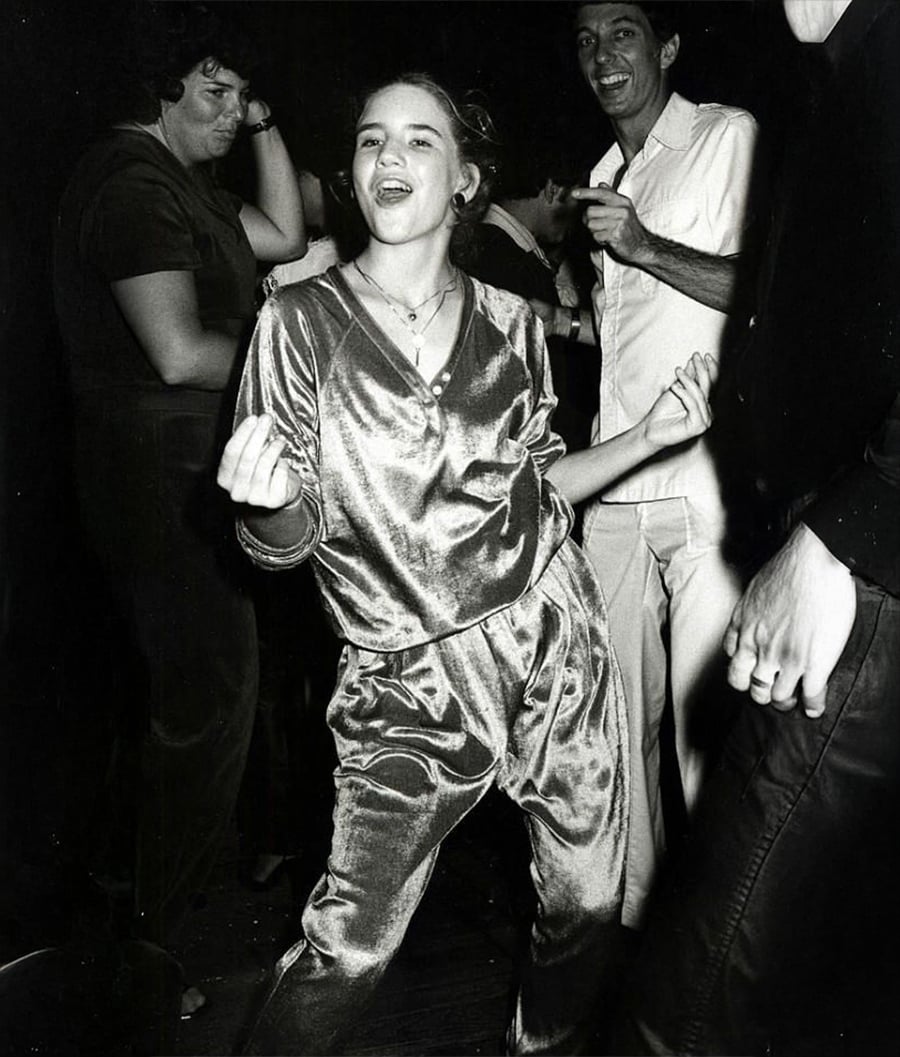 A young woman in a shiny, loose-fitting outfit dances energetically at a lively party, surrounded by people who are also enjoying themselves. The photo is black and white, capturing a joyful moment.