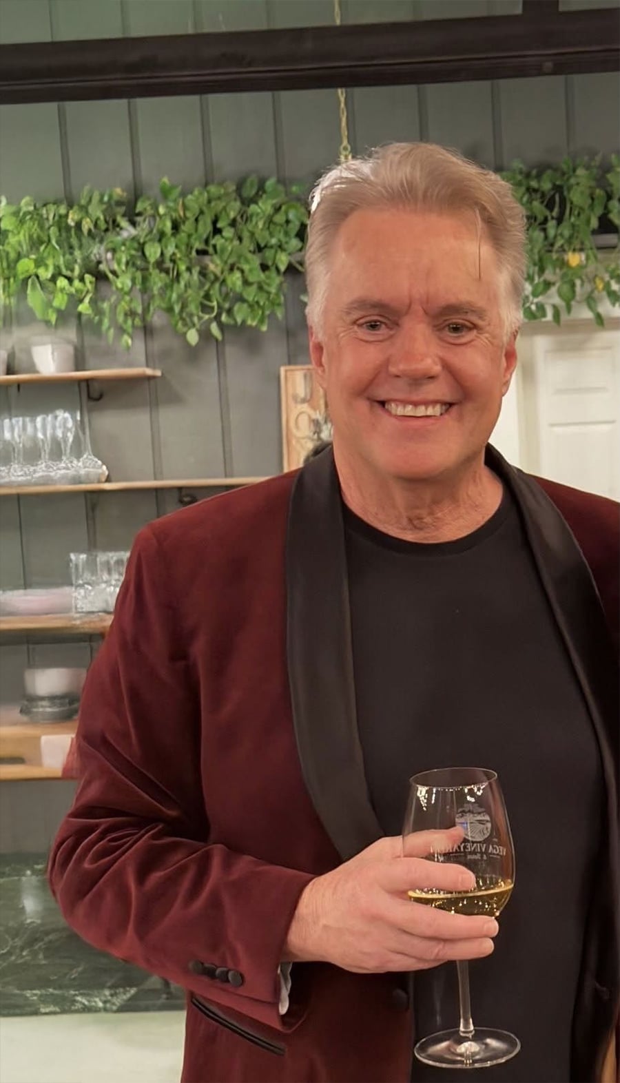 A smiling man in a maroon velvet jacket and black shirt holds a glass of white wine. Behind him are shelves with glasses and plants on a green wooden wall.
