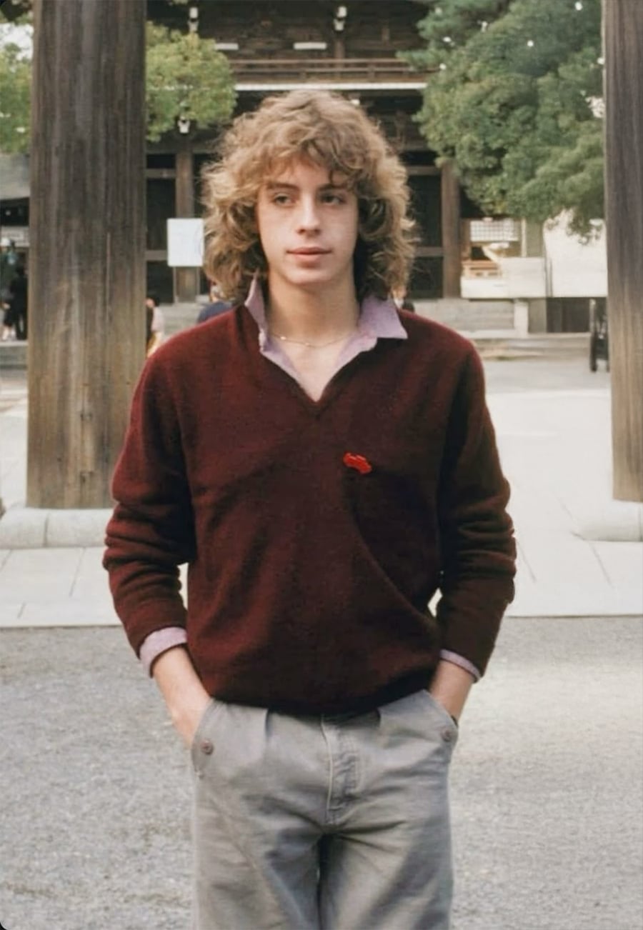 A young person with curly blond hair, wearing a maroon sweater over a collared shirt and light gray pants, stands outdoors with hands in pockets. Large wooden pillars and trees are visible in the background.