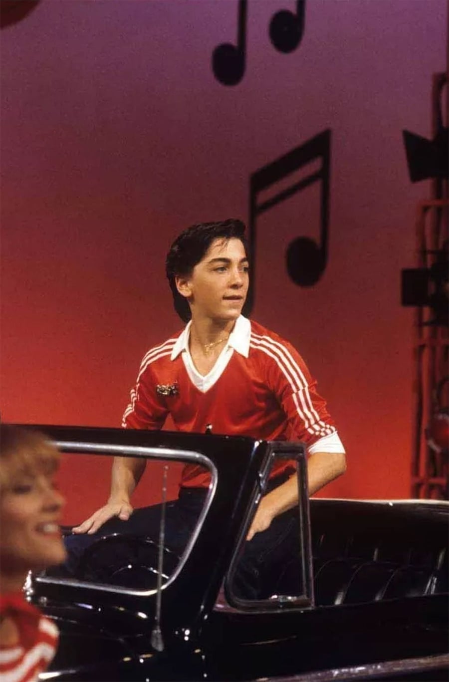 A young man in a red and white striped shirt sits on the back of a black convertible car on a stage with a red background and large black musical notes hanging as decorations.
