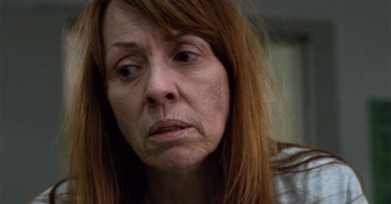 A middle-aged woman with long reddish-brown hair looks down with a sad, tired expression on her face. She is indoors and wearing a gray top. The lighting is subdued, and her surroundings are blurred in the background.