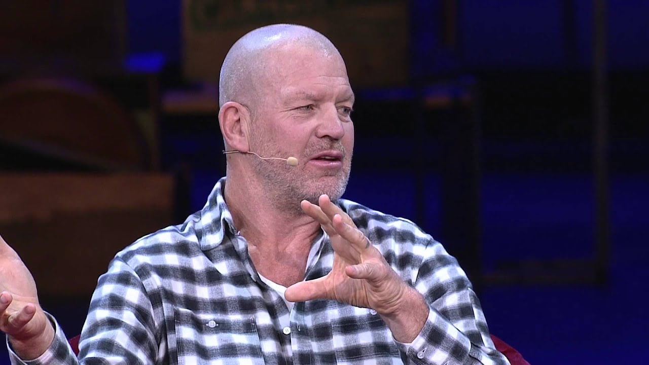 A bald man wearing a black-and-white plaid shirt speaks animatedly on stage, gesturing with his hands and using a headset microphone. The background is dimly lit.