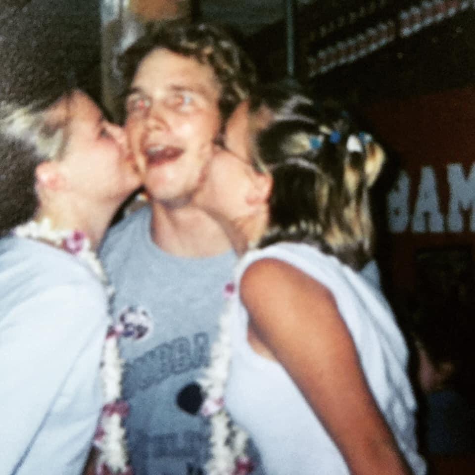 A young man wearing a gray t-shirt smiles widely as two young women, each wearing leis, kiss him on the cheeks. The background is slightly blurred with indistinct signs and decorations.