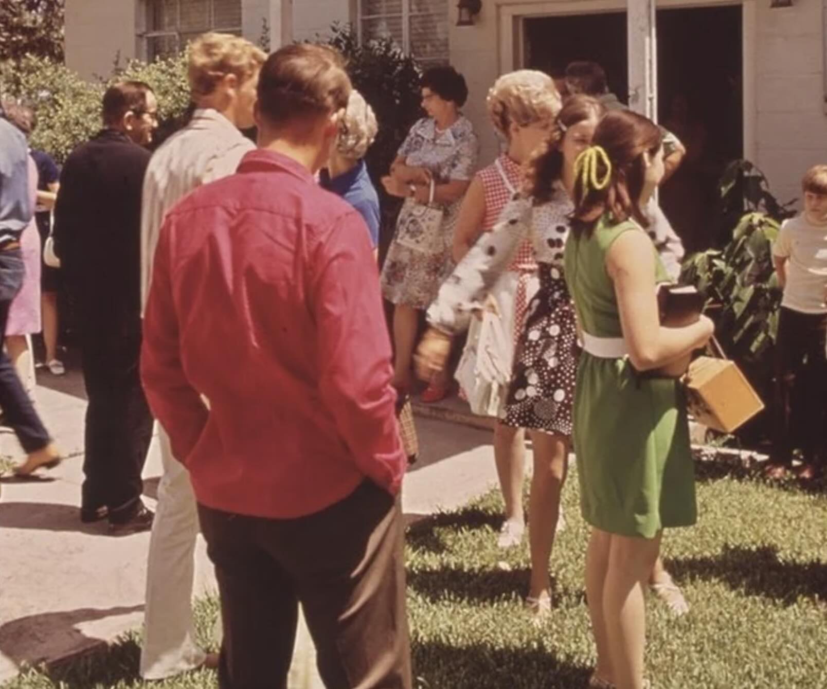 A group of people, mostly adults, socialize outside a house on a sunny day. One girl in a green dress with a yellow ribbon in her hair holds books and a box. Others wear casual and patterned clothes.