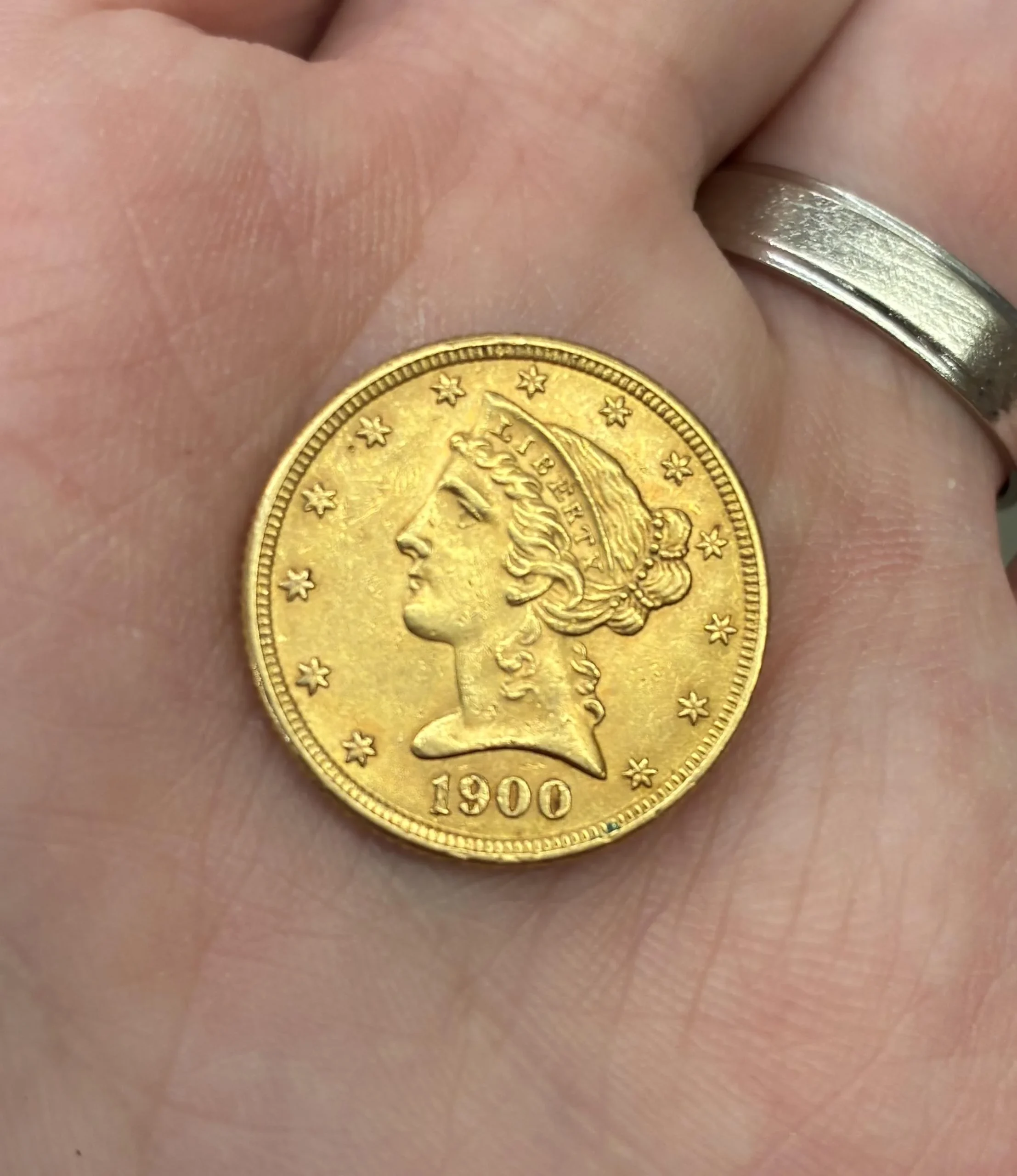 A hand holding a gold coin featuring the profile of Lady Liberty, surrounded by stars and the year "1900" inscribed at the bottom. A silver ring is visible on one finger.