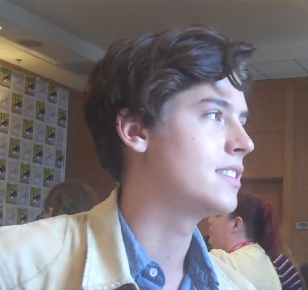 A young person with wavy brown hair, wearing a light-colored jacket over a blue shirt, is photographed in profile indoors with others in the background and a Comic-Con logo wall behind them.