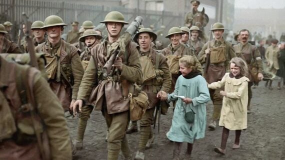 A group of World War I soldiers in uniform and helmets march through a muddy street, smiling. Two young girls in dresses run alongside them, holding hands and appearing cheerful. Buildings and fences are in the background.
