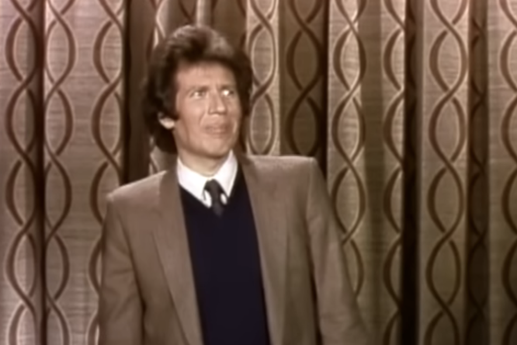 A man in a brown suit jacket, dark sweater, and tie stands in front of patterned curtains, looking upward with a puzzled expression.