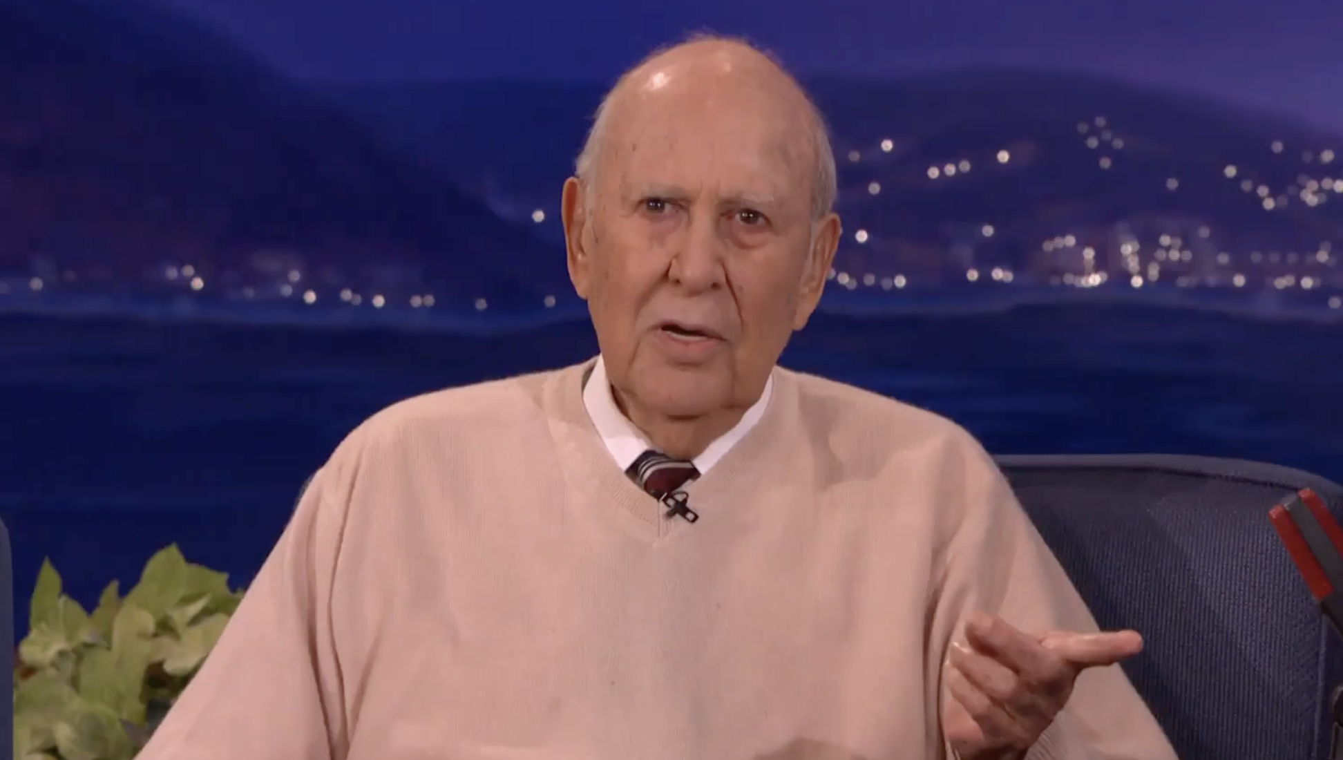 An older man with a bald head, wearing a beige sweater over a shirt and tie, sits in a studio setting with blurred lights and mountains in the background, gesturing with his right hand while speaking.