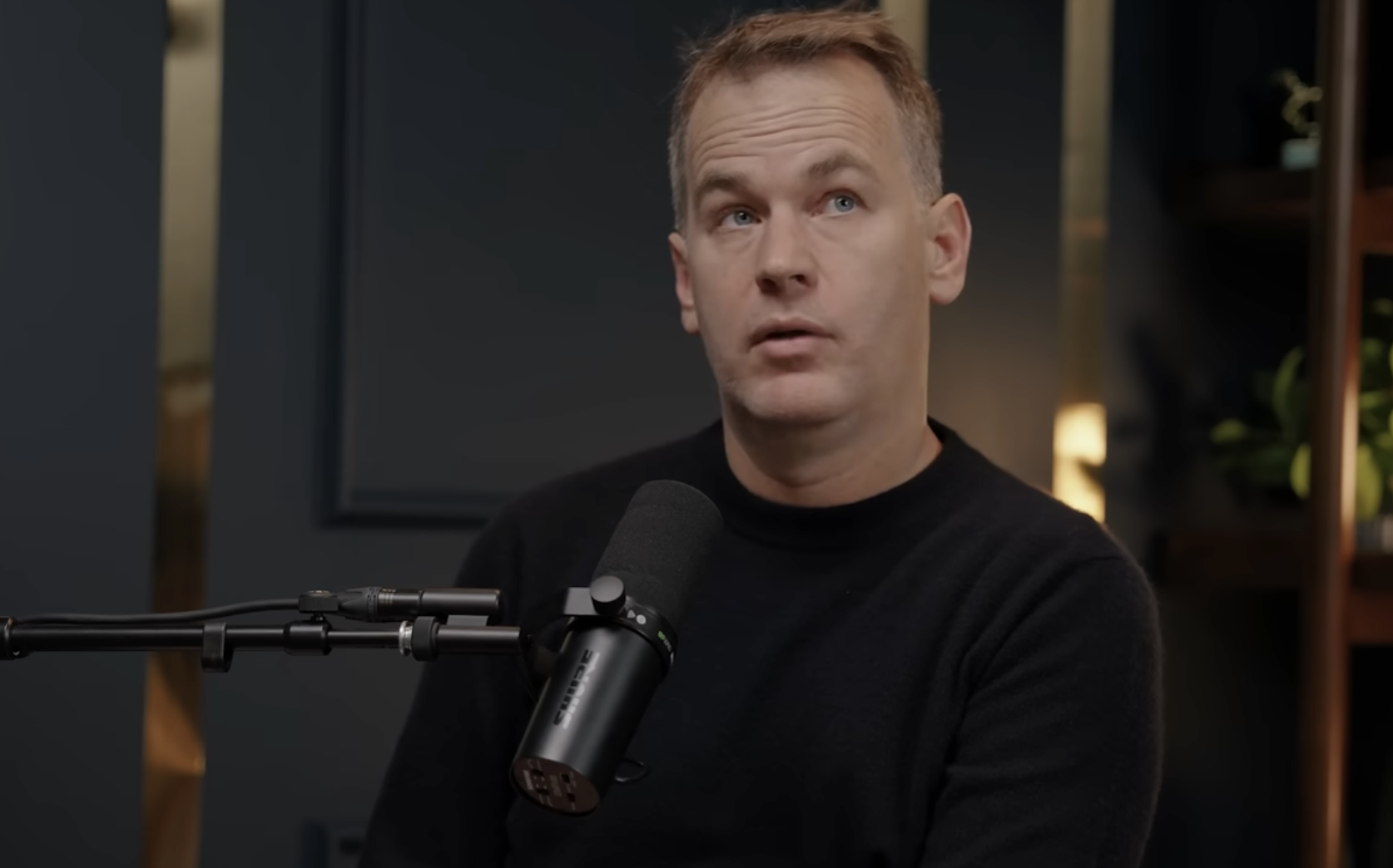 A man in a black sweater sits in front of a microphone, looking slightly upward. The background includes dark walls and some plants.