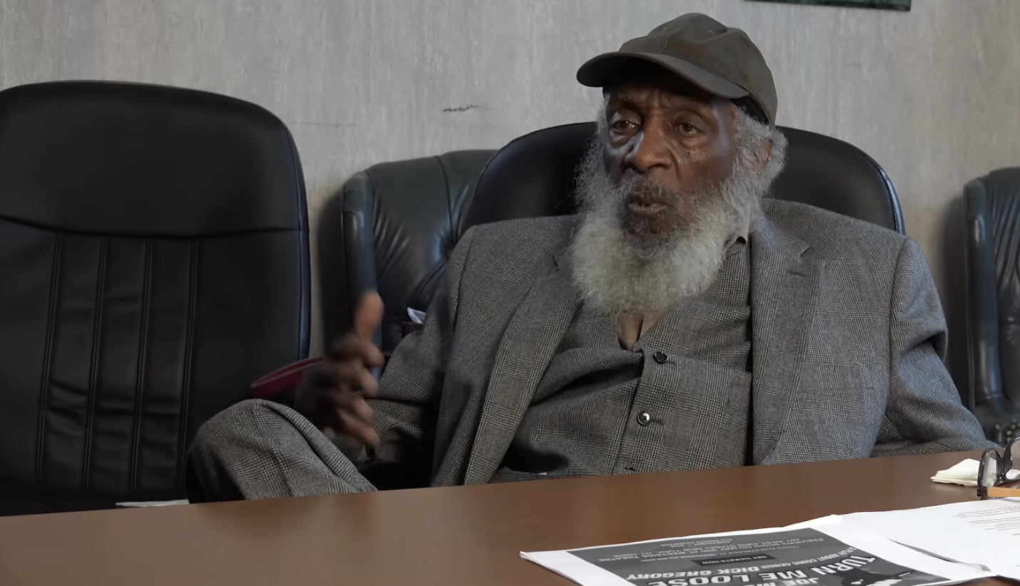 An older man with a gray beard and cap sits at a conference table, gesturing with one hand. He is wearing a gray suit, and there are papers and glasses on the table in front of him.