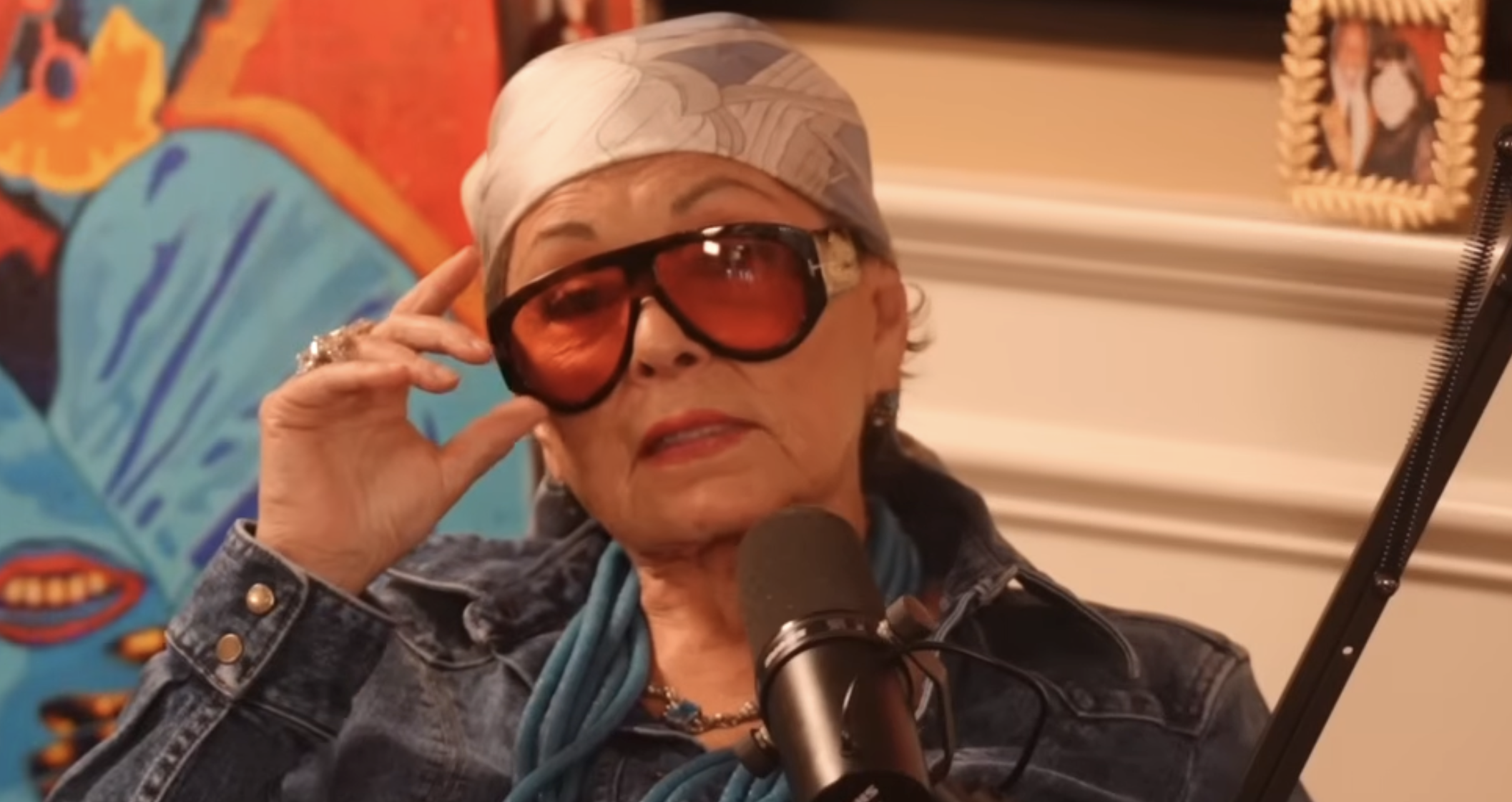A woman wearing large tinted sunglasses, a patterned headscarf, and a denim jacket sits in front of a microphone, with colorful artwork and a framed photo visible in the background.