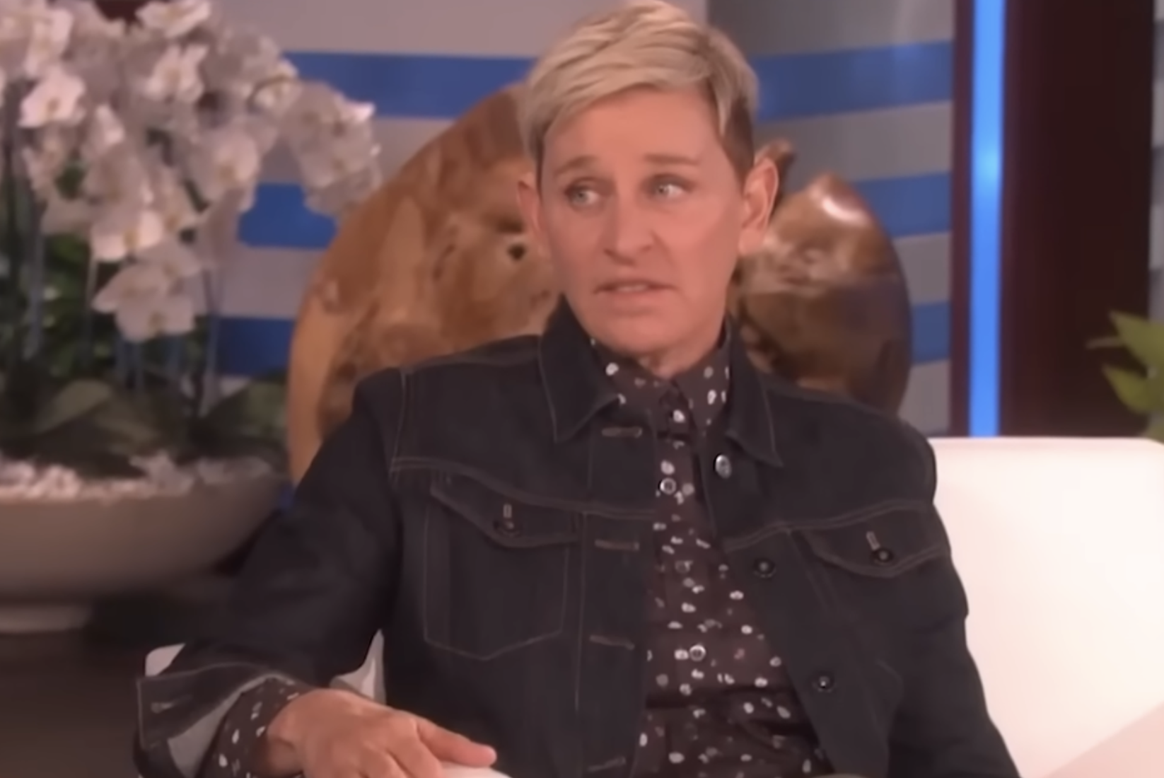 A person with short blond hair, wearing a dark denim jacket and patterned shirt, sits on a white couch in a studio setting with flowers and a wooden decoration in the background.