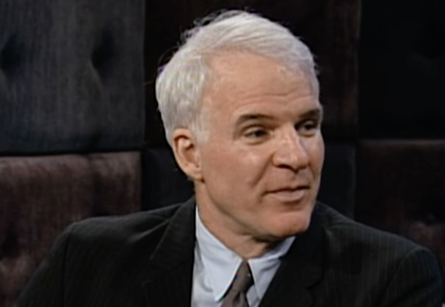 A man with short white hair, wearing a dark suit and light blue shirt, is seated and looking to his right. The background features dark, tufted upholstered panels.