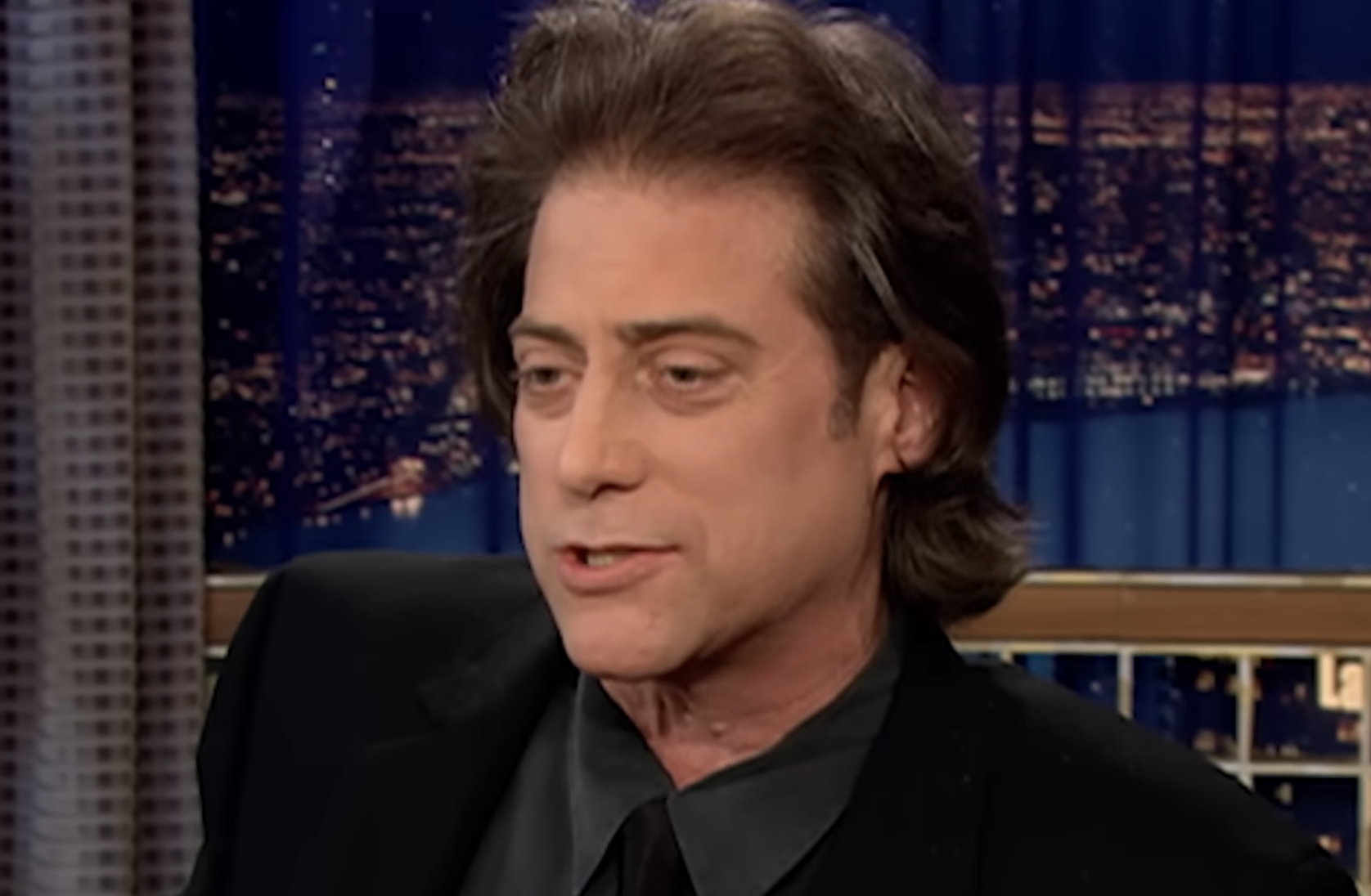 A man with medium-length brown hair wearing a black jacket and shirt sits indoors, speaking. A cityscape at night is visible through the window behind him.