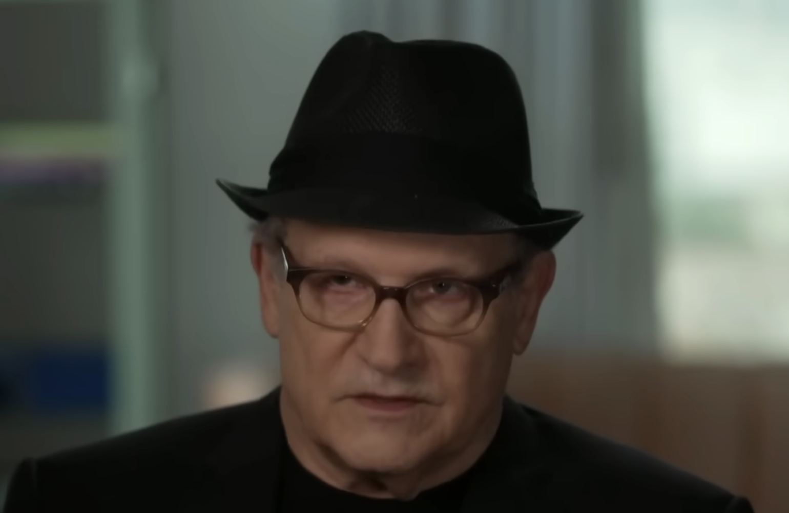 A person wearing a black fedora hat, glasses, and a black shirt is sitting indoors with a blurred background. The lighting is soft and the expression appears serious.