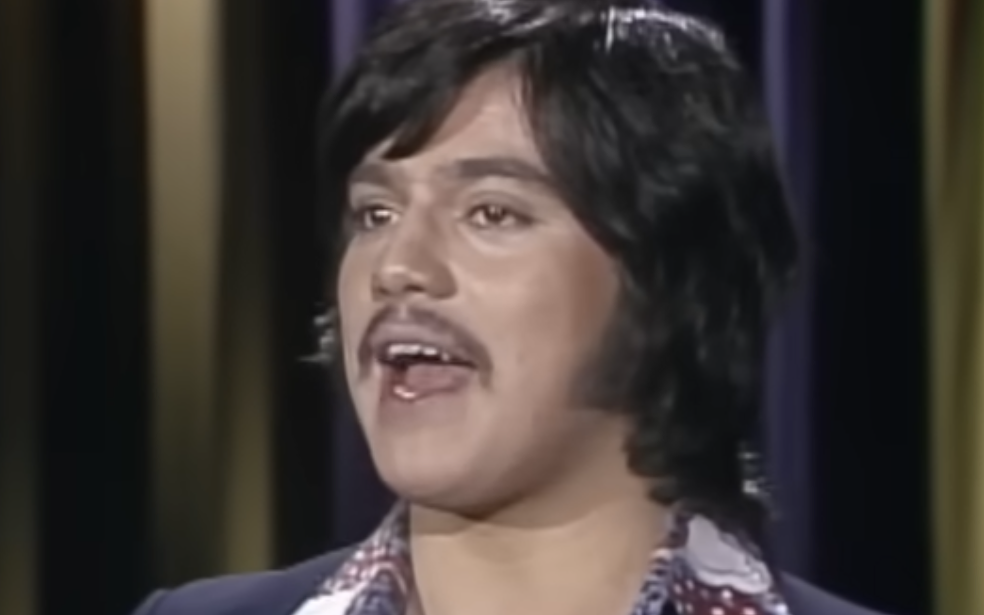 A person with medium-length dark hair, a mustache, and sideburns is singing or speaking with their mouth open, wearing a dark jacket over a patterned shirt, in front of a background with vertical stripes.