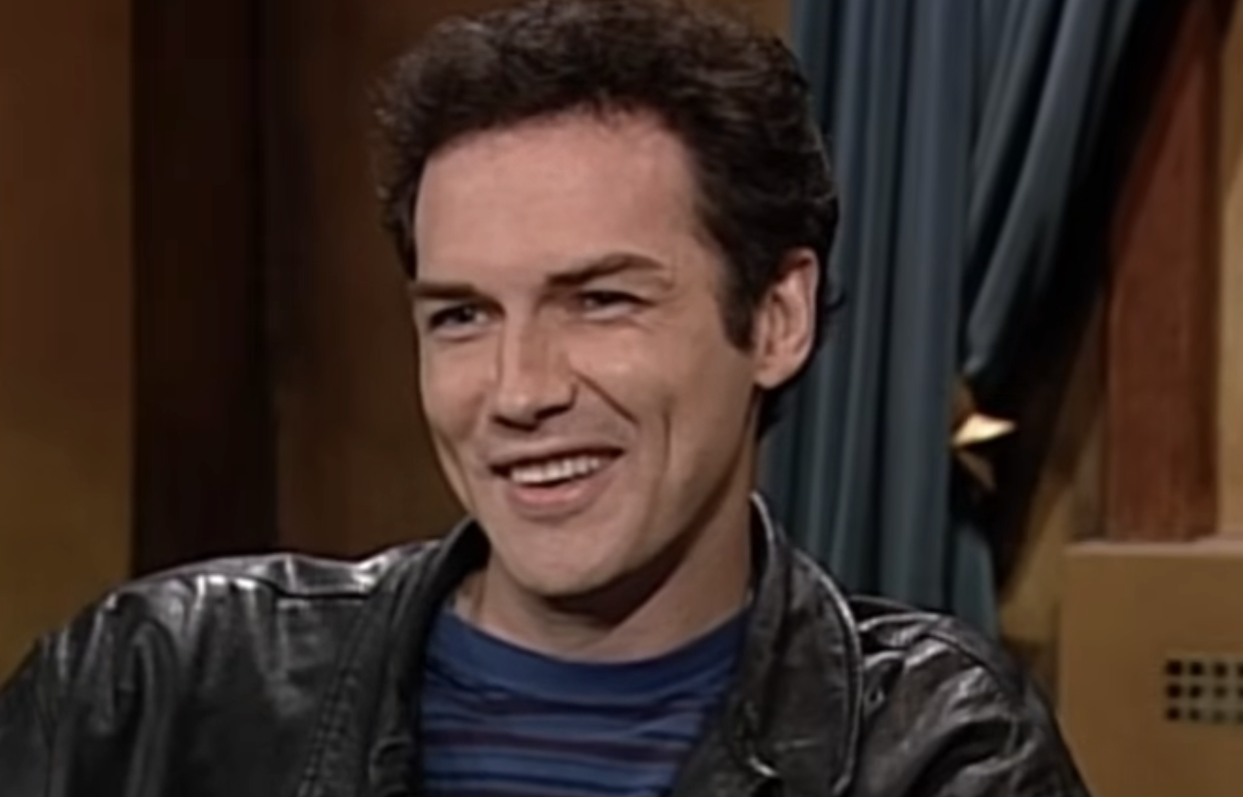 A man with short dark hair is smiling while sitting indoors. He is wearing a black leather jacket over a blue and black striped shirt. There is a curtain and part of a wall visible in the background.