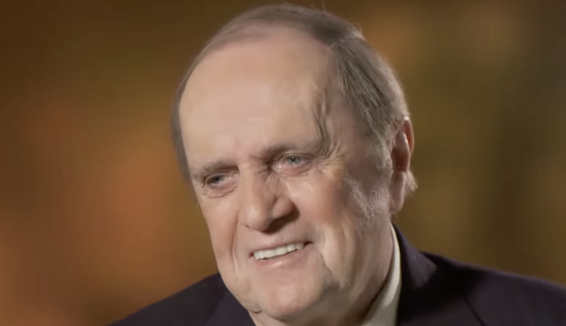 An older man with short, thinning hair and a slight smile, wearing a dark suit jacket and light shirt, is pictured against a softly blurred, warm-toned background.