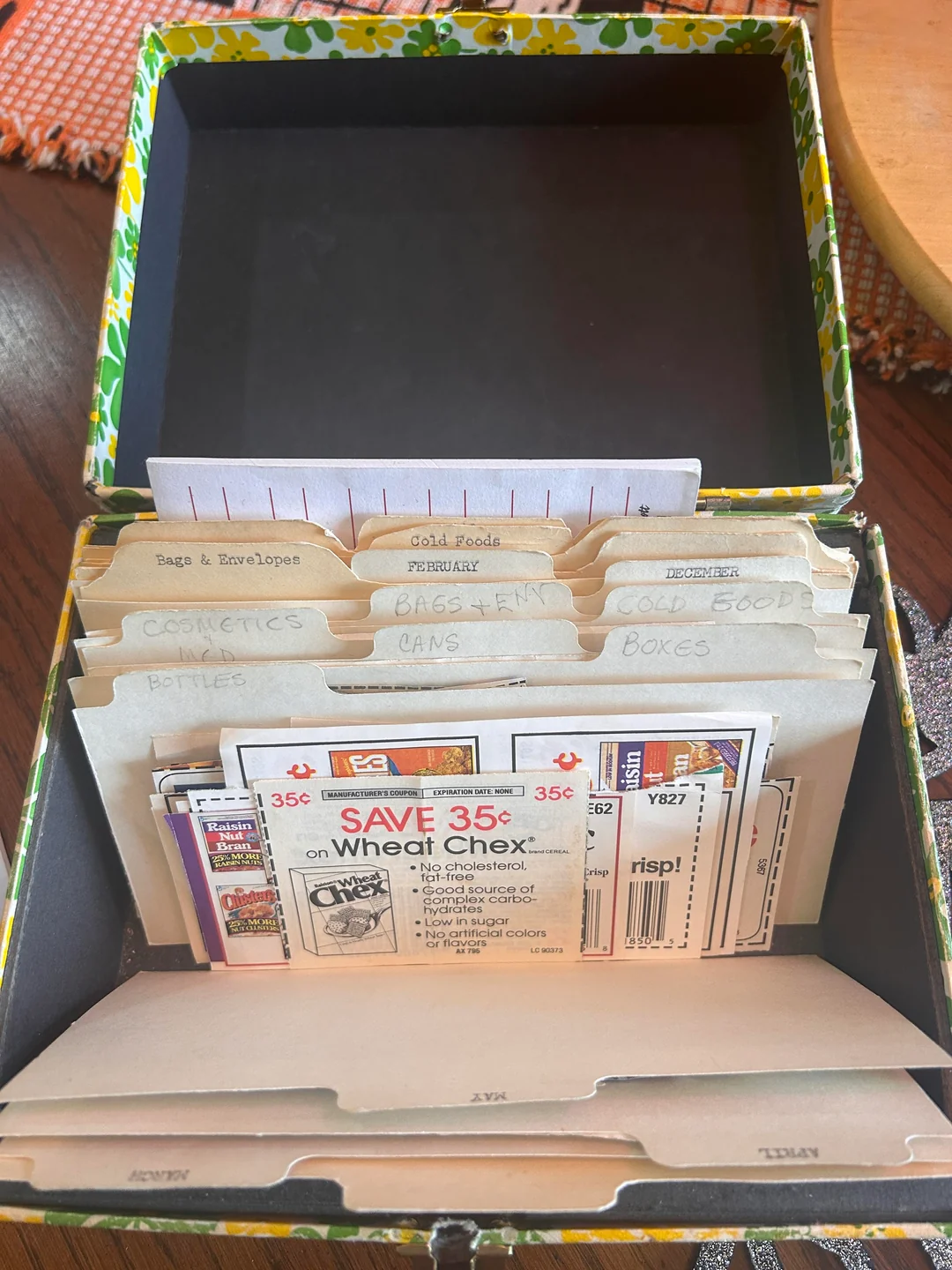 A recipe box with labeled dividers such as “COSMETICS,” “CANS,” and “GOLD FOOD” organizes paper coupons, including one for 35¢ off Wheat Chex, inside a floral-patterned container on a wooden table.