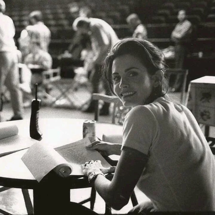 A woman sits at a round table, smiling at the camera while holding papers. People and filming equipment are visible in the blurred background, suggesting a busy set or rehearsal environment. The photo is in black and white.