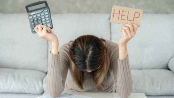 A person sits at a table with head bowed, holding a calculator in one hand and a cardboard sign reading "HELP" in the other, suggesting financial stress or difficulty.