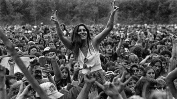 A black-and-white photo of a large outdoor crowd at a concert or festival. A smiling woman sits on someone&rsquo;s shoulders in the center, raising both hands in peace signs. Many people surround her, cheering and raising their arms.