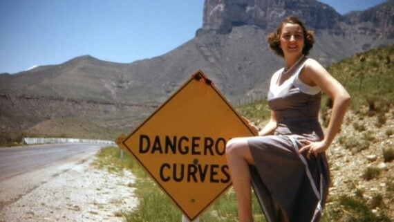 A woman in a gray dress poses playfully with one leg raised on a yellow road sign that reads "DANGEROUS CURVES," beside a rural road with mountains in the background under a clear blue sky.