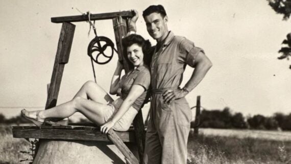 A smiling woman sits playfully on a wooden well structure while a man stands beside her, both dressed in casual vintage clothing, posing outdoors in a grassy field.